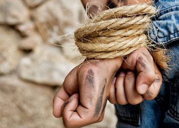 Dirty prisoner, arms bound behind back with rope, standing next to a stone wall.